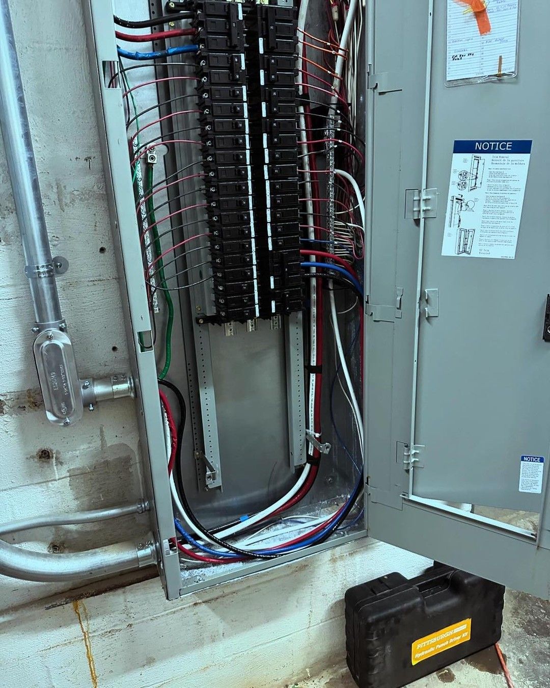 Open electrical panel with wires and circuit breakers. Gray metal box mounted on a concrete wall.