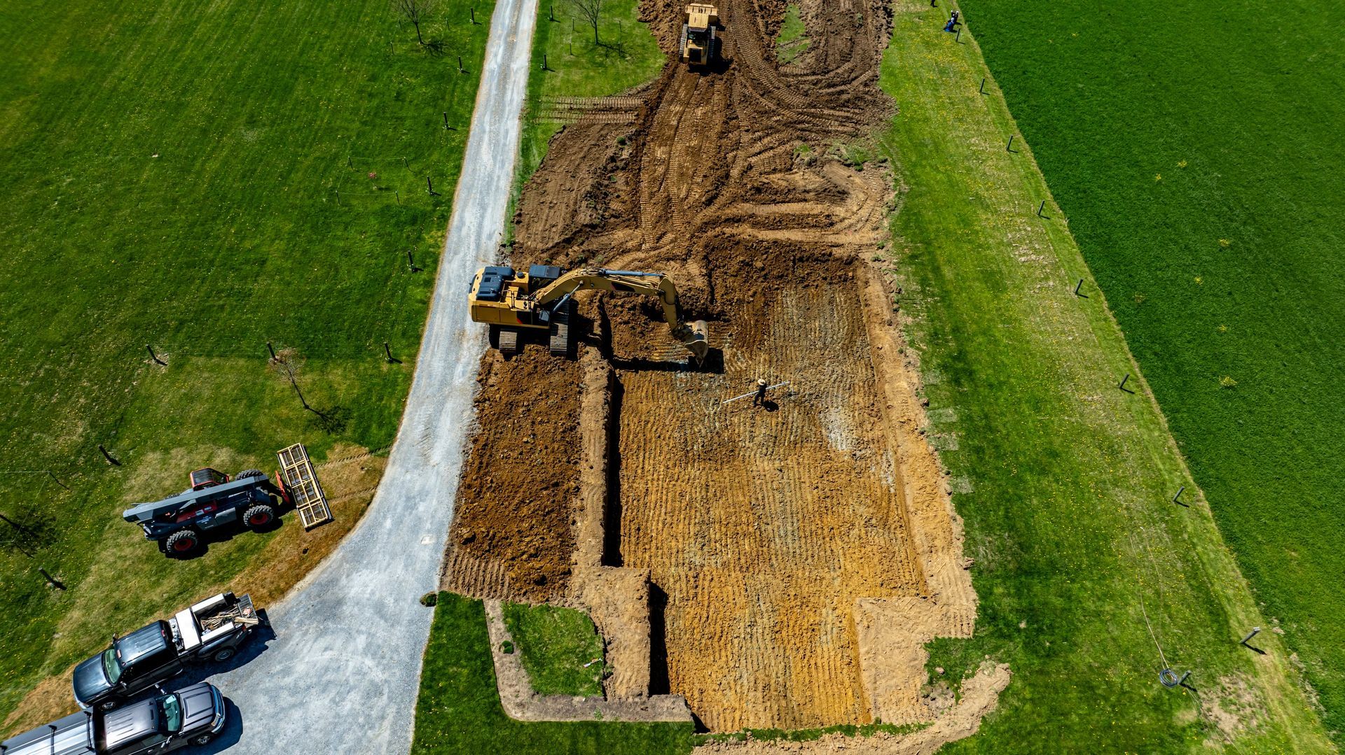Aerial view of road construction with excavators digging a long dirt trench beside a paved road and green fields.