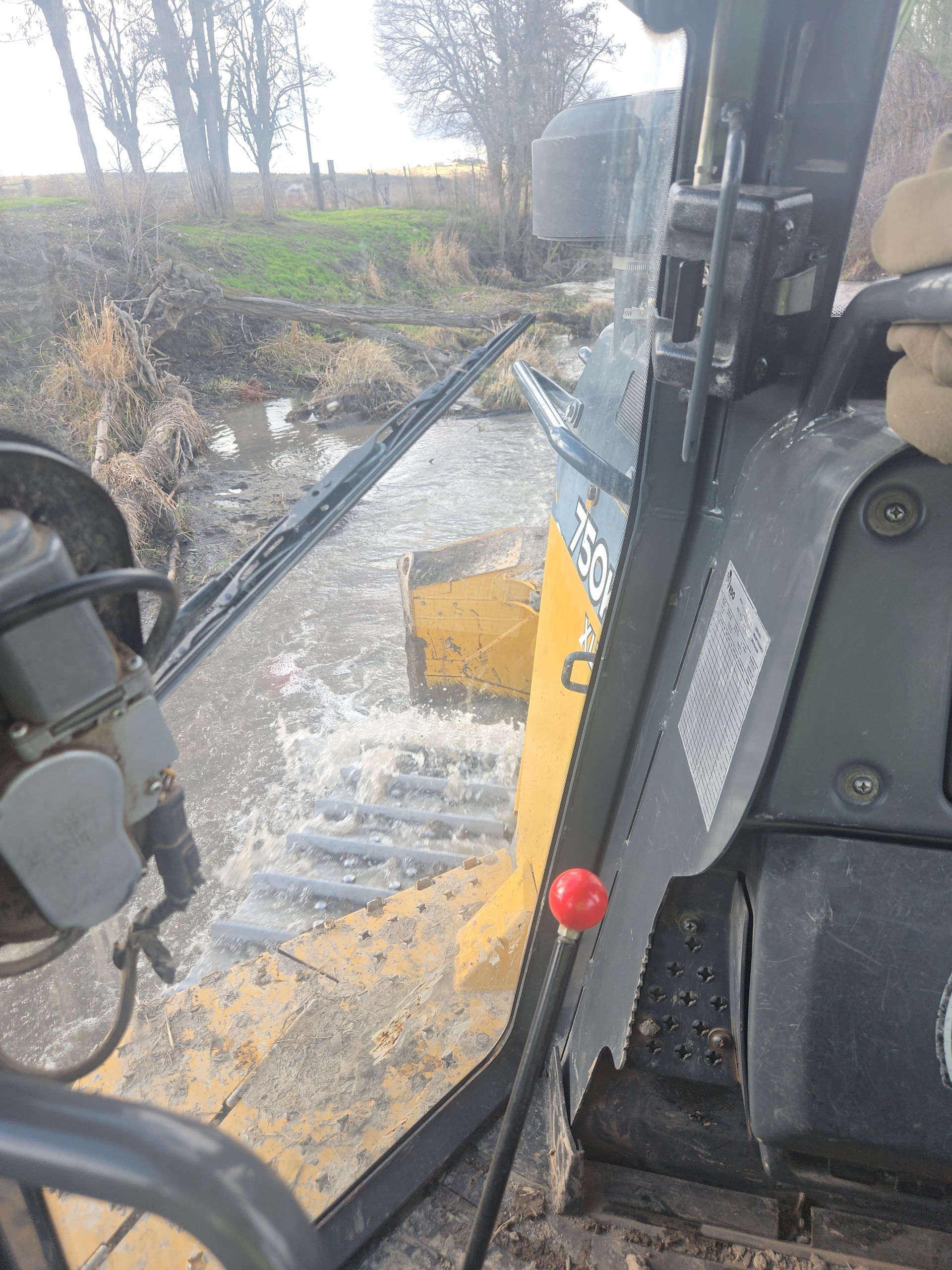 Inside a construction vehicle cab, muddy yellow exterior and controls visible beside a wet roadside.
