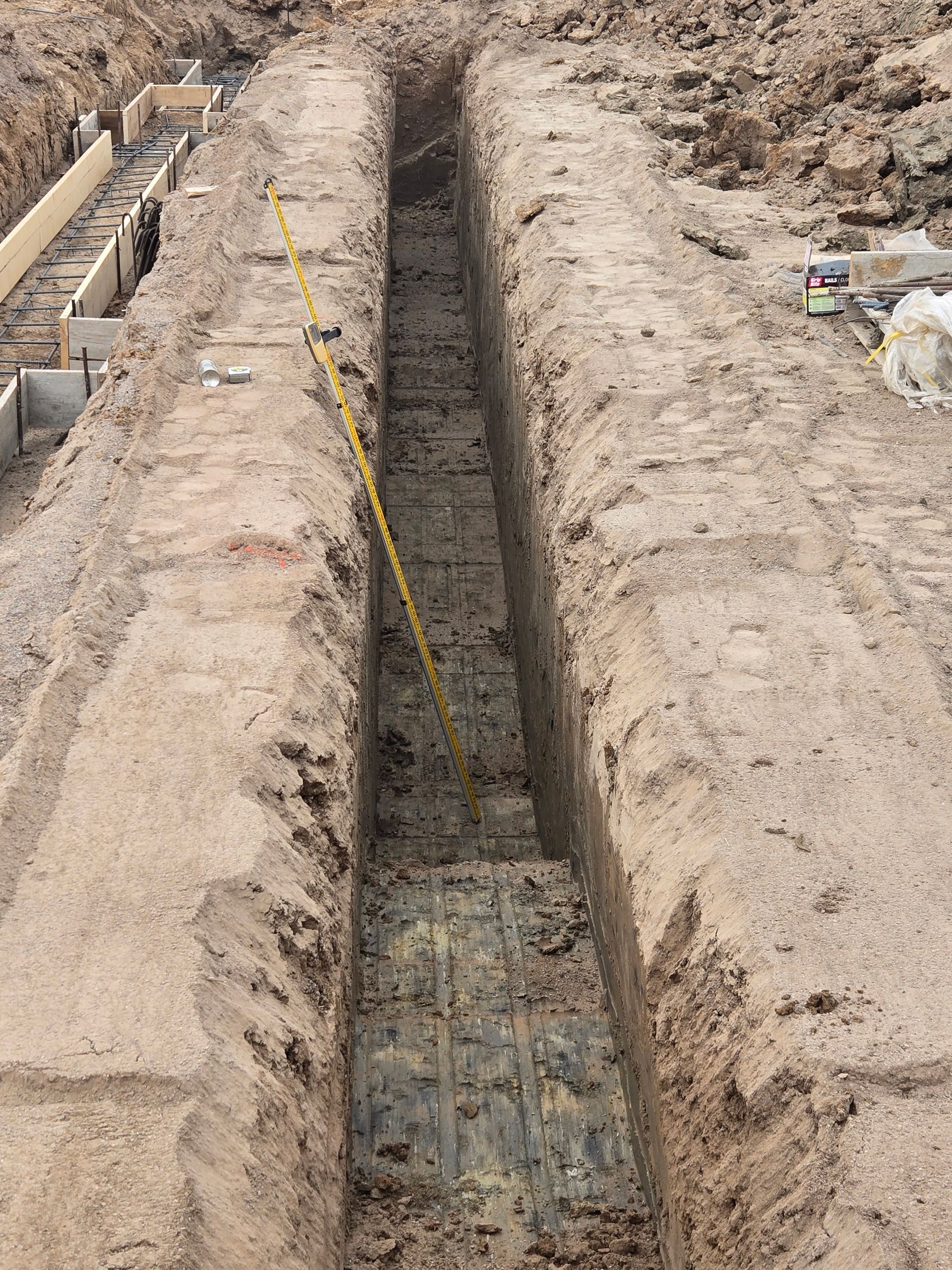 Narrow trench dug in dirt at a construction site, with exposed concrete at the bottom and caution tape along the edge