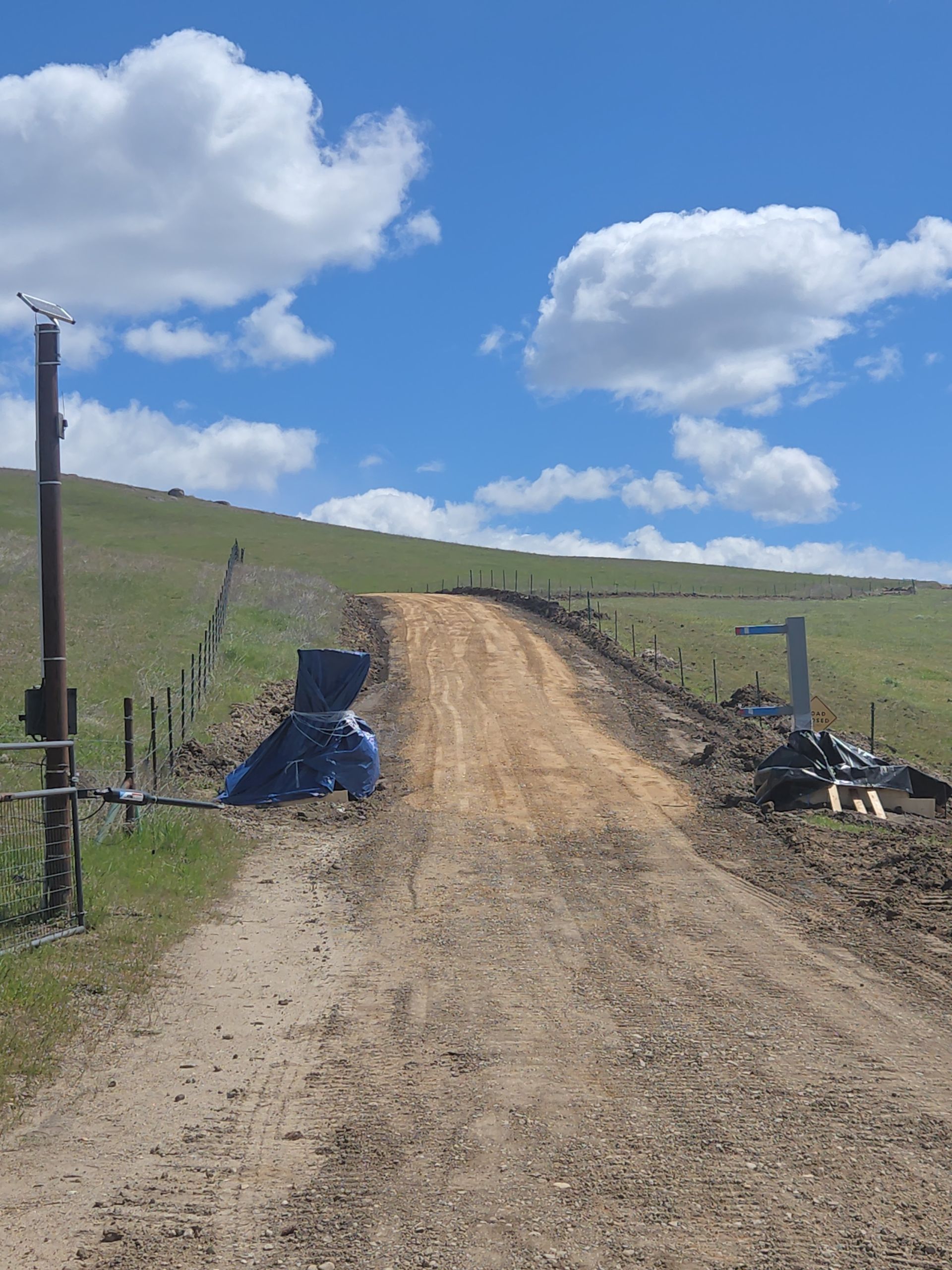 Dirt road uphill through grassy hills under a blue sky with clouds, bordered by fence posts and a gate.