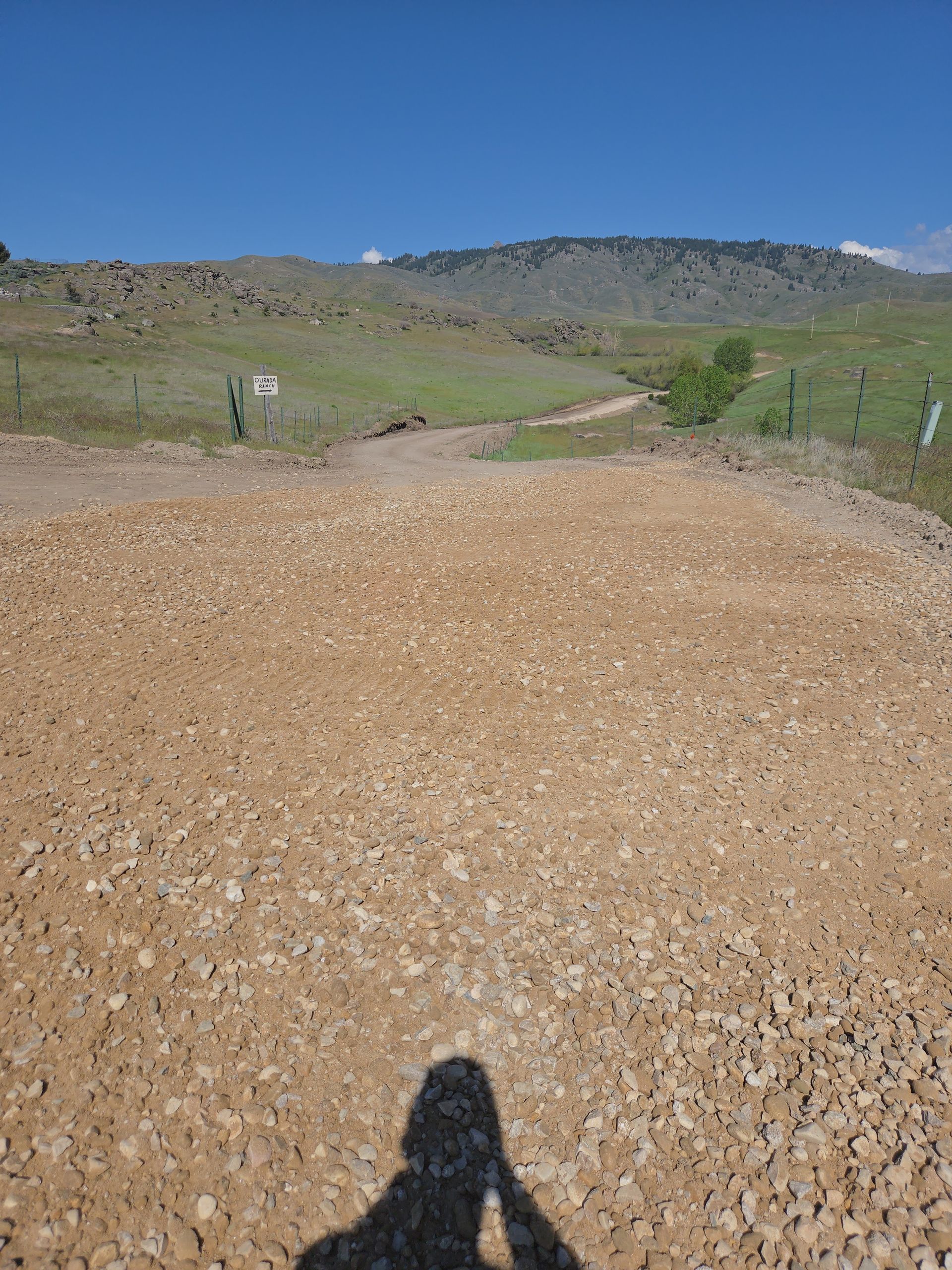 Rocky dirt road through green hills under a blue sky, with a person’s shadow in the foreground