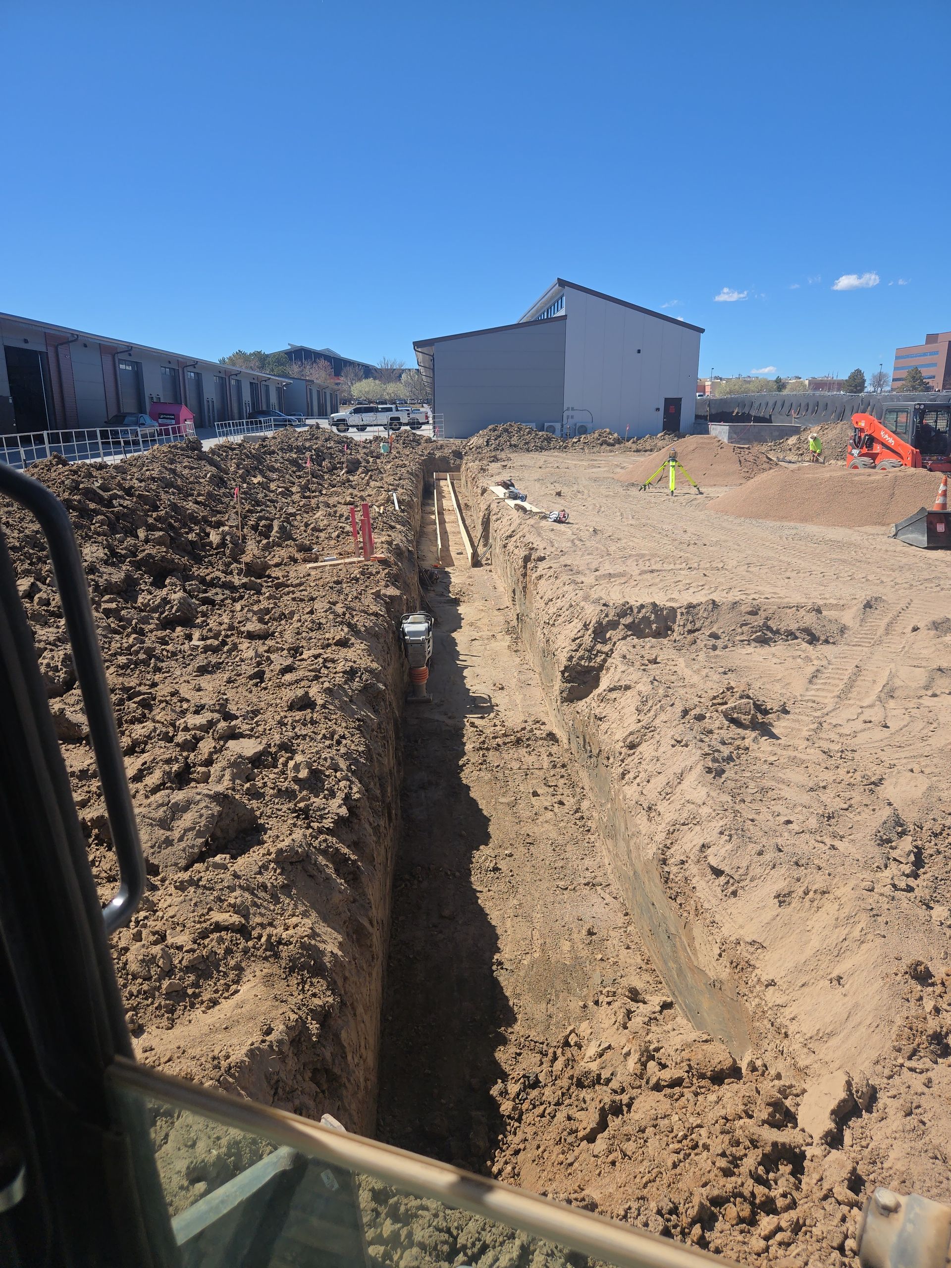 Construction trench beside a gravel road, with houses and parked vehicles under a clear blue sky.