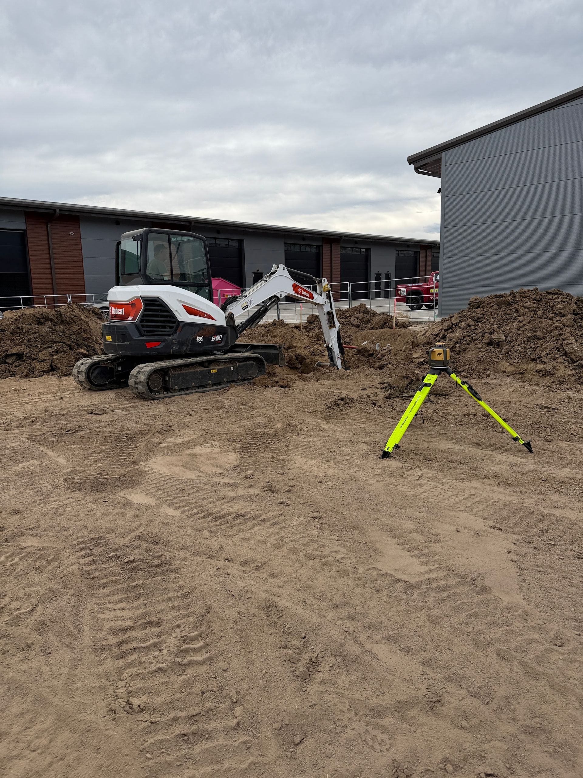 Construction site with a small white excavator, dirt piles, and a yellow tripod on muddy ground.