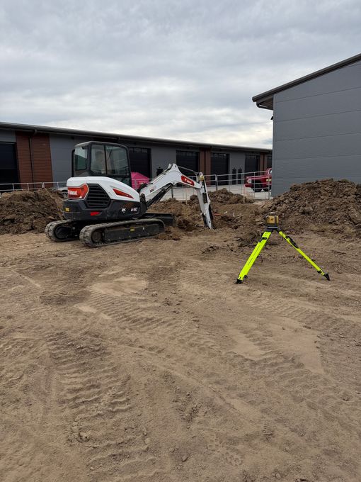 Construction site with a small white excavator, dirt piles, and a yellow tripod on muddy ground.