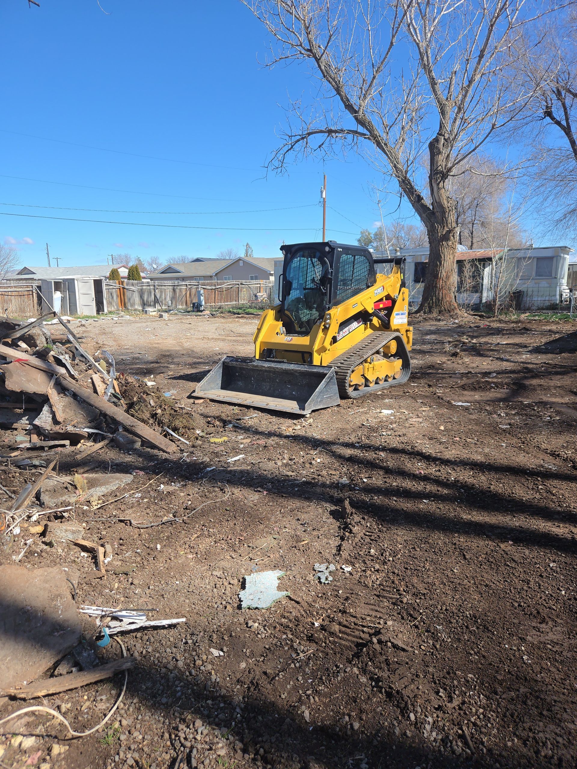Yellow skid-steer loader clearing debris in a dirt lot under a blue sky
