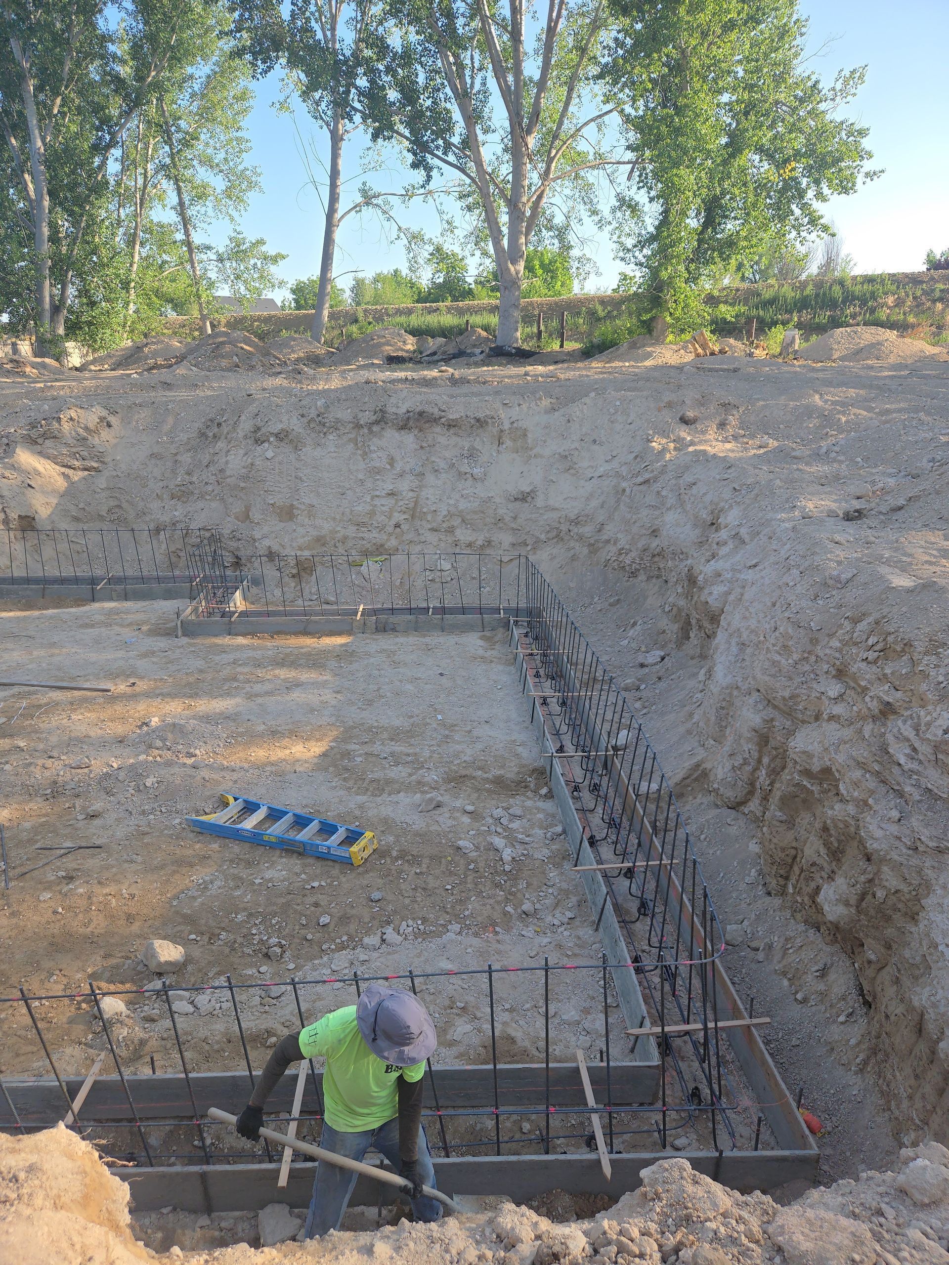 Worker in green shirt at a concrete foundation excavation with rebar and trees above.