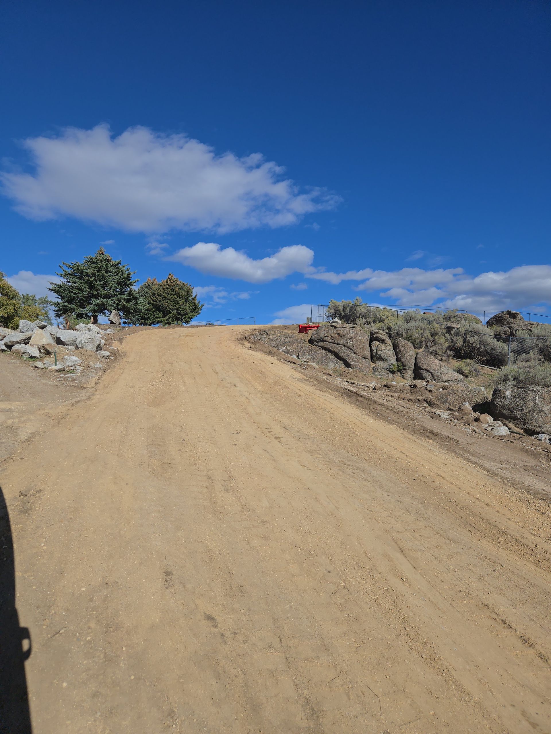 Dirt road climbing a hill under a bright blue sky with scattered clouds and sparse shrubs.