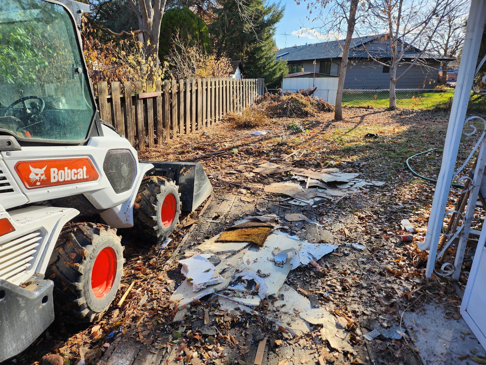 Bobcat skid steer in a muddy backyard beside debris, fence, and fallen leaves