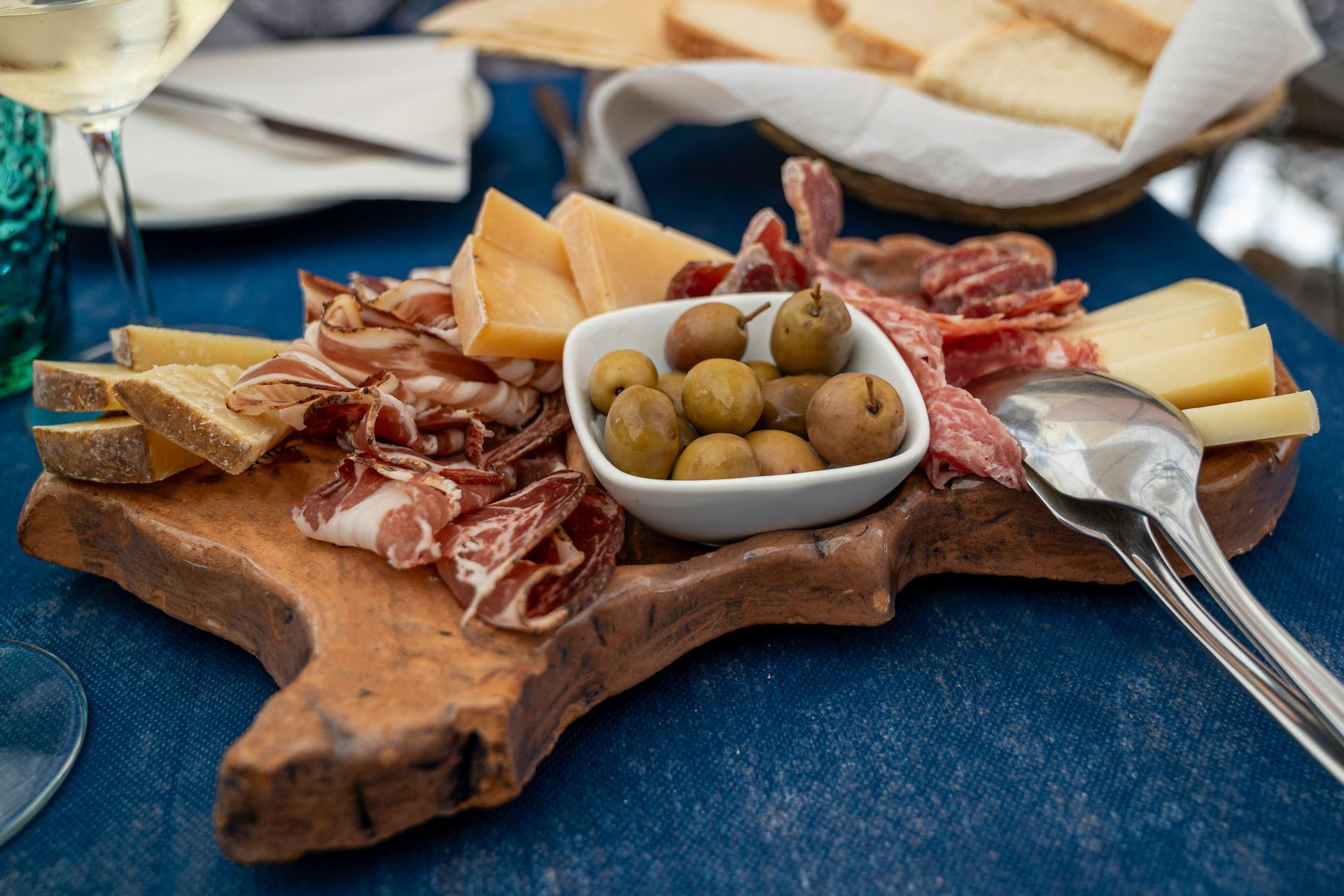 Tagliere di salumi con carni, formaggi e olive su una tovaglia blu; pane sullo sfondo.