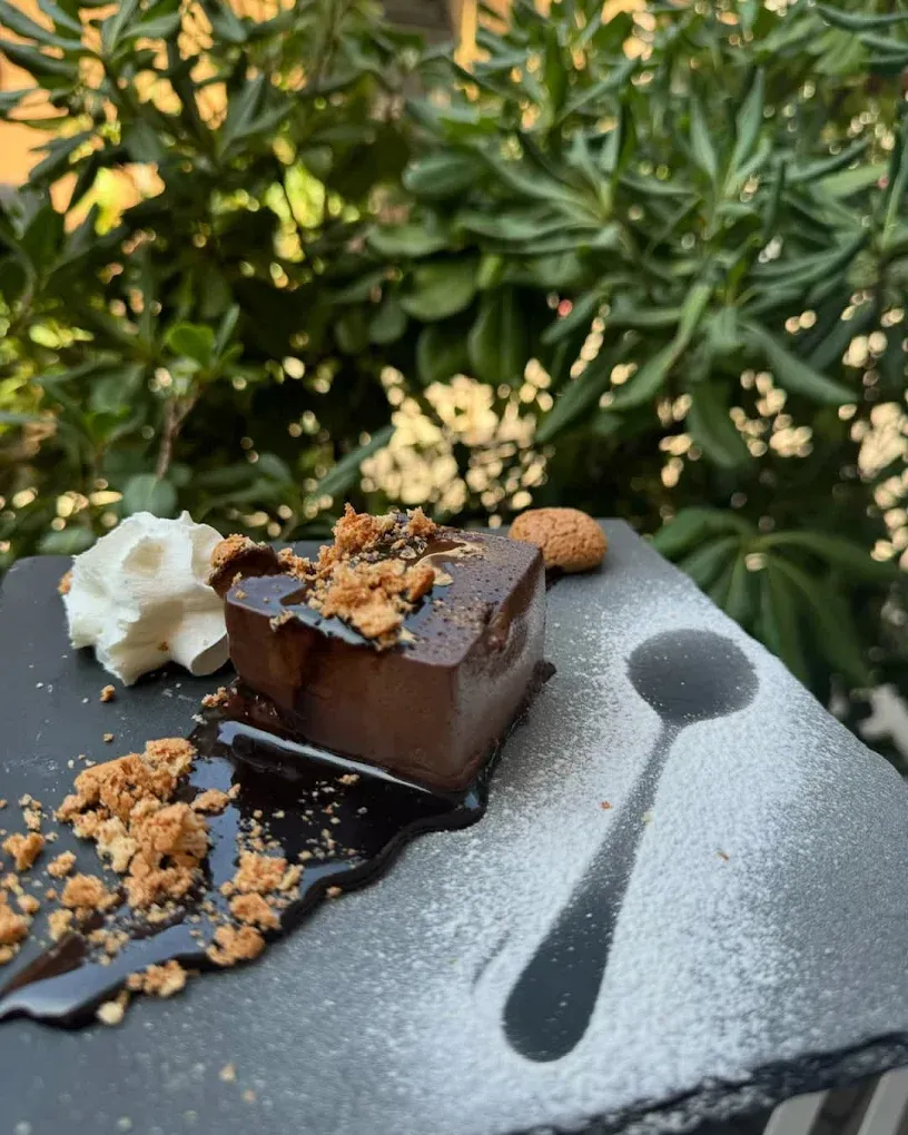 Dolce al cioccolato servito su un piatto di ardesia nera con panna montata, briciole di biscotti e un cucchiaino di zucchero a velo.