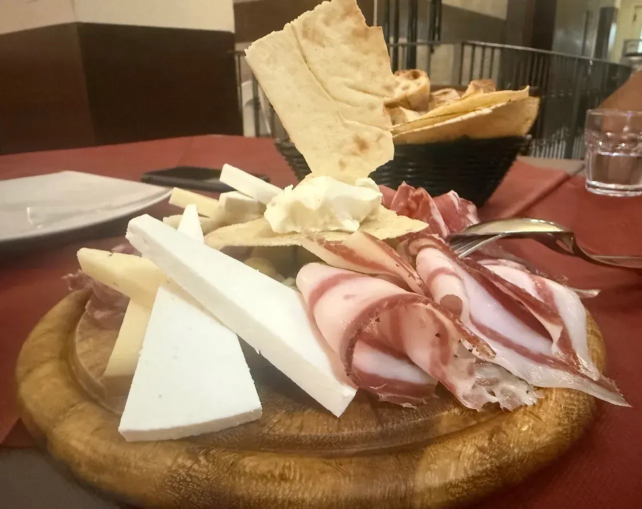 Vassoio di legno con formaggi assortiti, salumi e pane, su una tovaglia rossa.