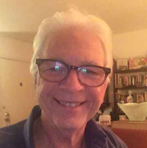 A person with light hair and glasses smiles while looking toward the camera in a room with a bookshelf in the background.