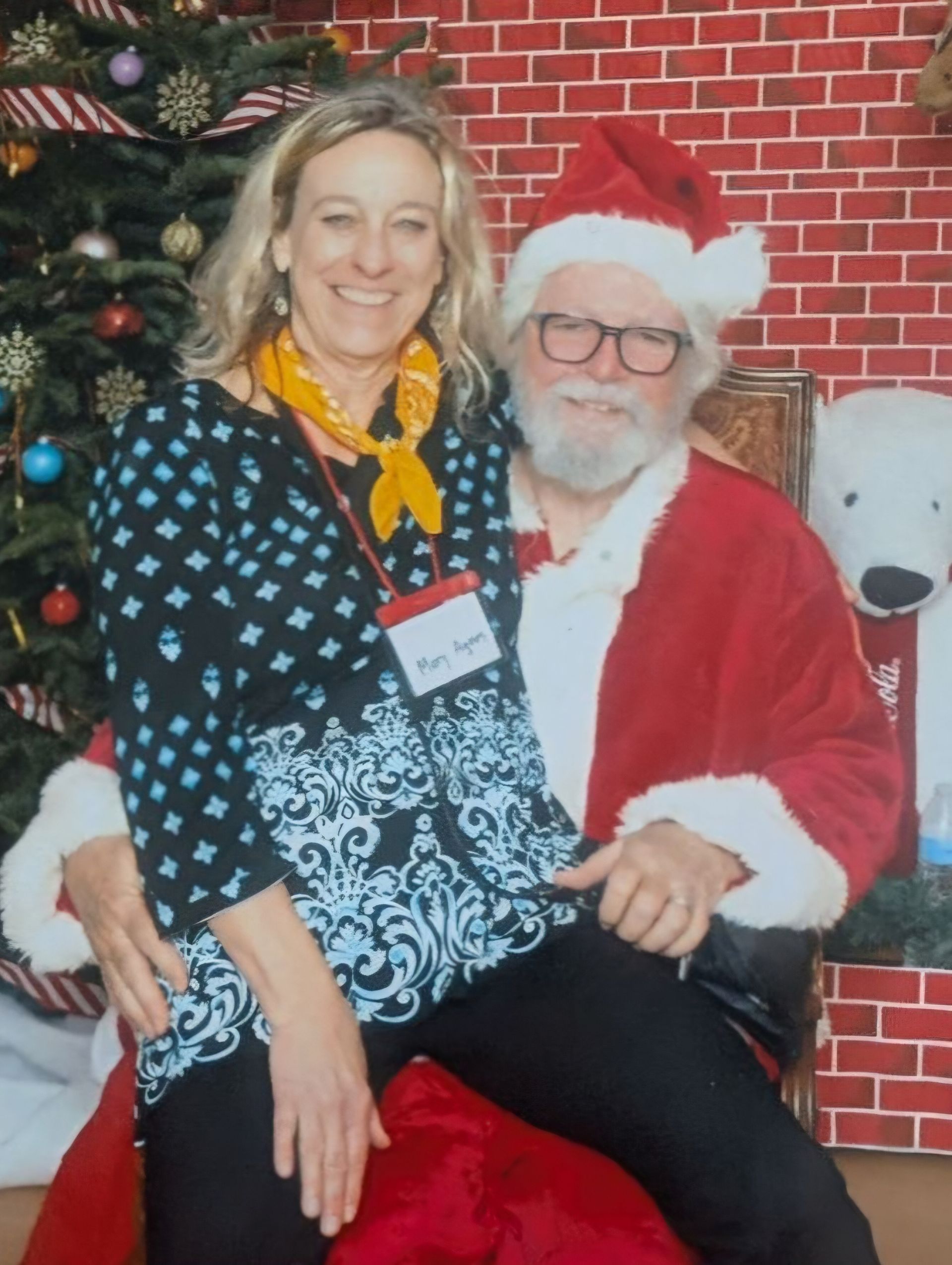 A smiling person sits on Santa Claus's lap in front of a Christmas tree and brick-patterned backdrop.