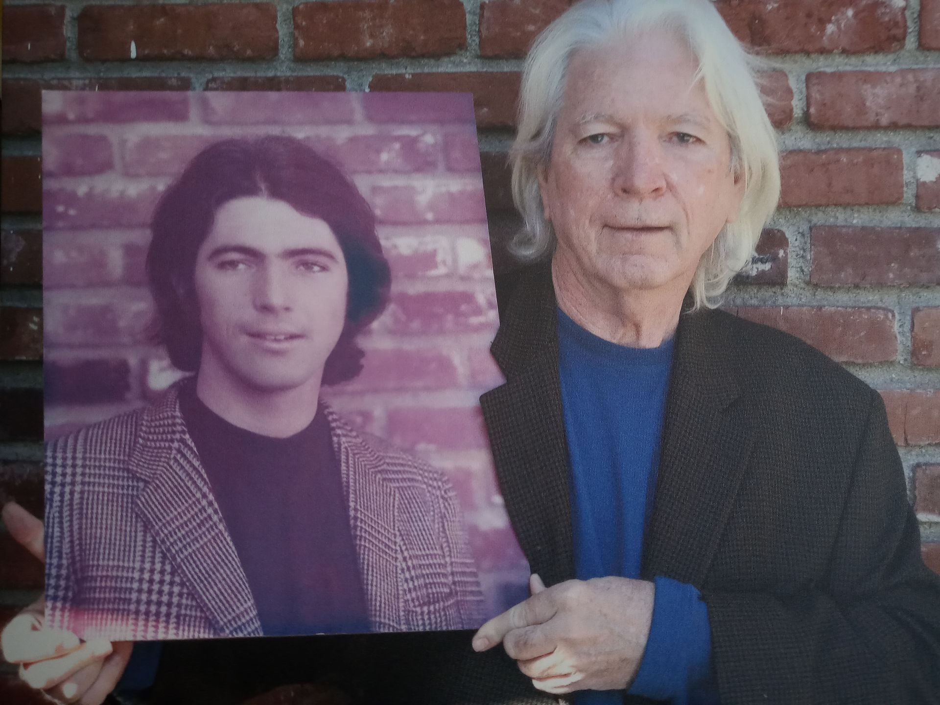 An older person holding a vintage photograph of a younger person, both wearing similar plaid jackets against a brick wall.