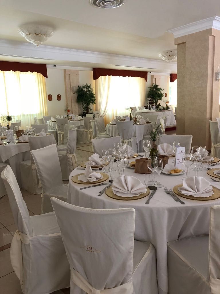 Una sala per banchetti allestita con tavoli rotondi bianchi, sedie coperte e tovaglie per un evento formale.