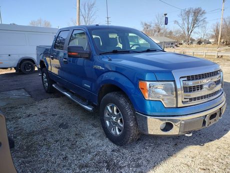 Blue Ford pickup truck parked on a gravel lot beside a white van, under clear daylight.