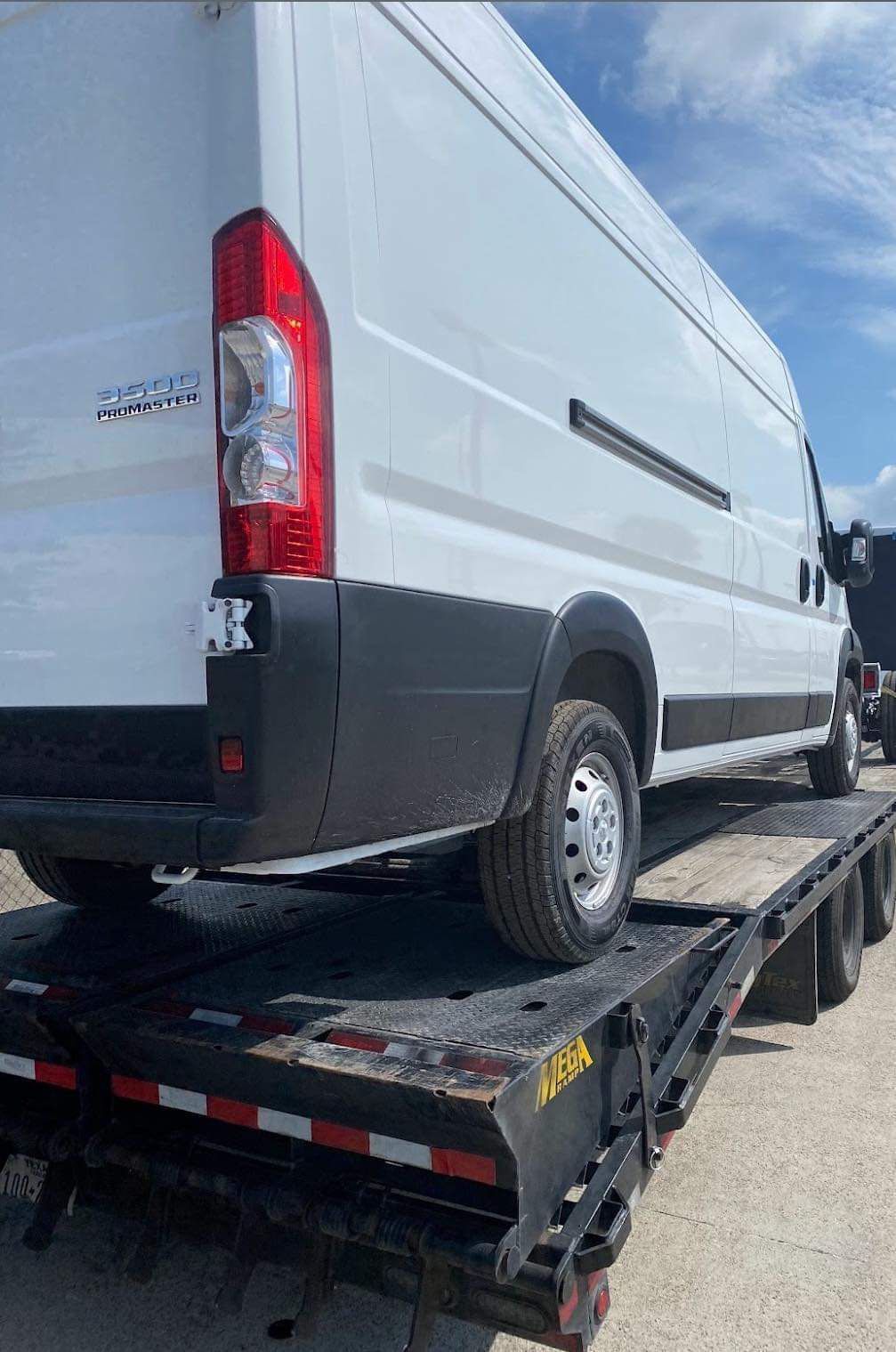 White cargo van strapped on a black flatbed trailer under a partly cloudy sky.