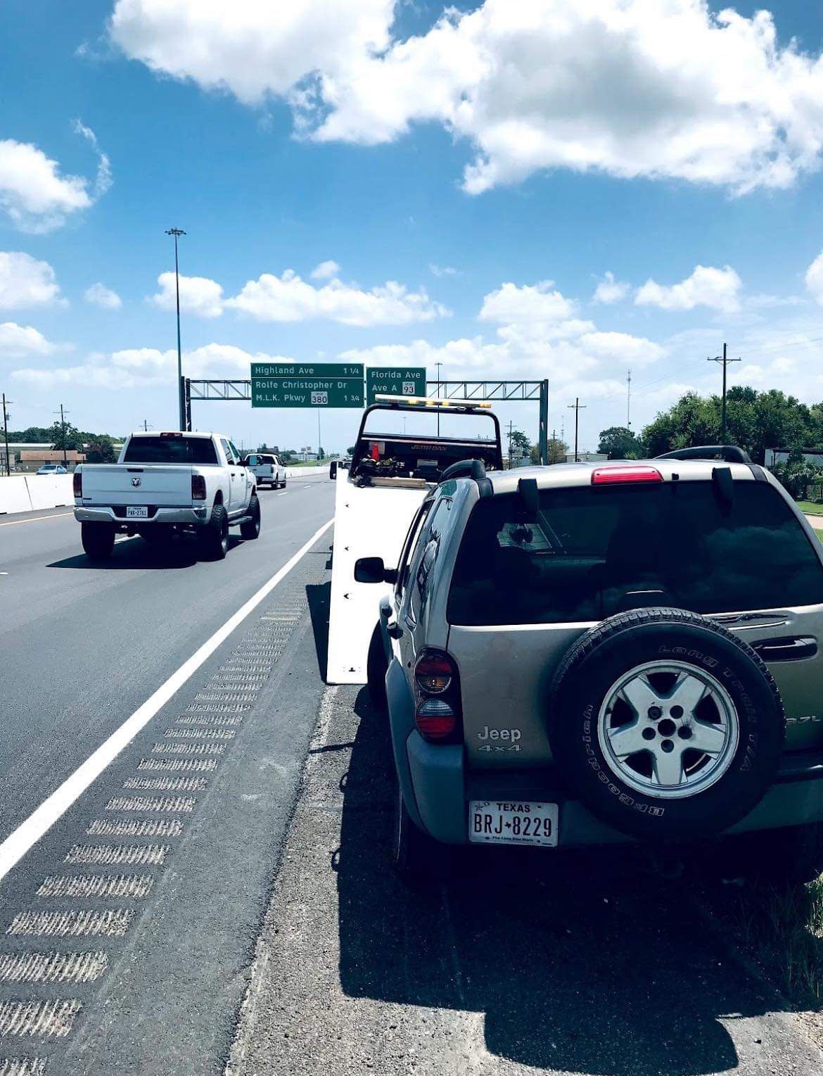 SUVs and a tow truck pulled over on a sunny highway shoulder, with green road signs ahead.
