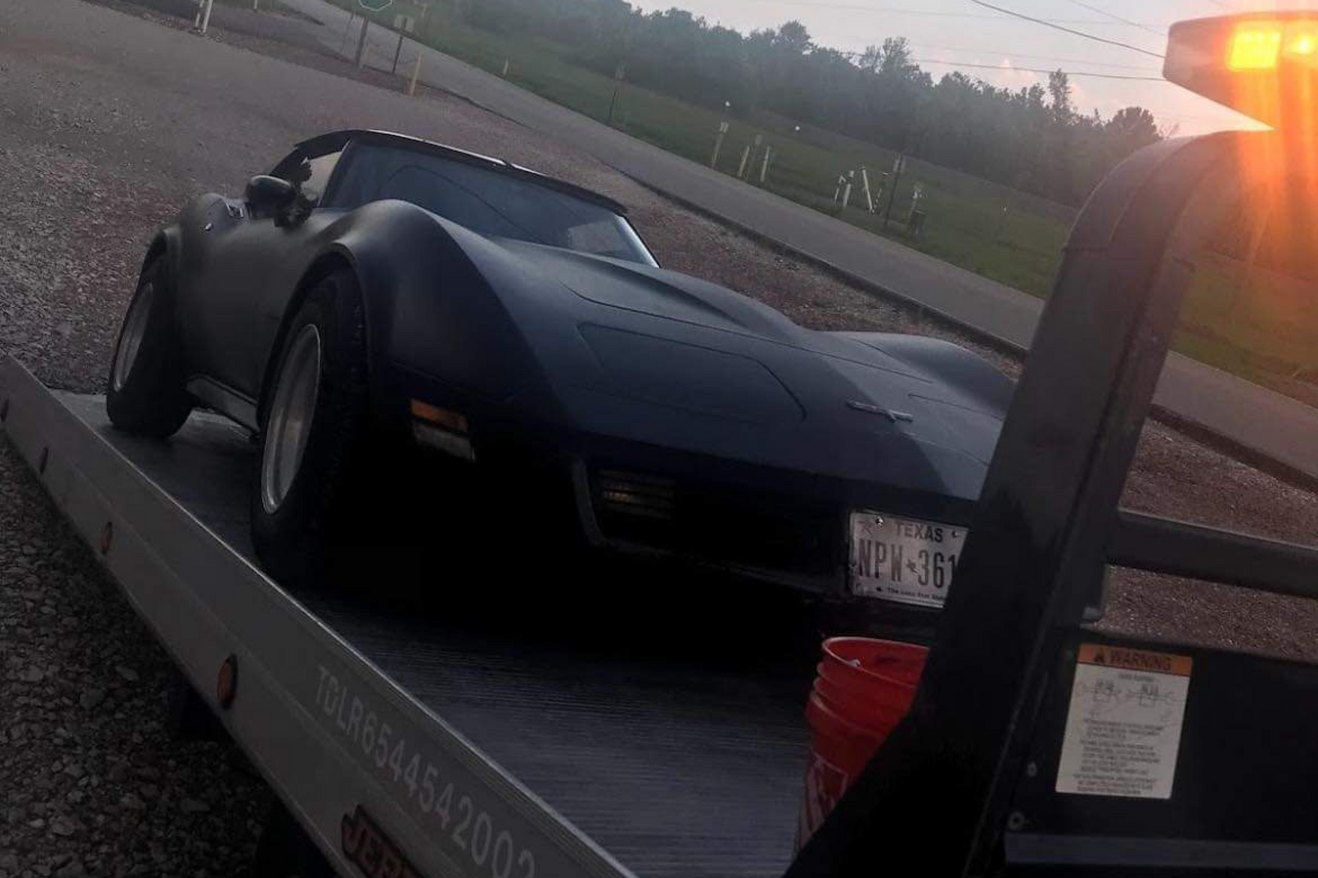 Black sports car on a flatbed tow truck at sunset beside a road