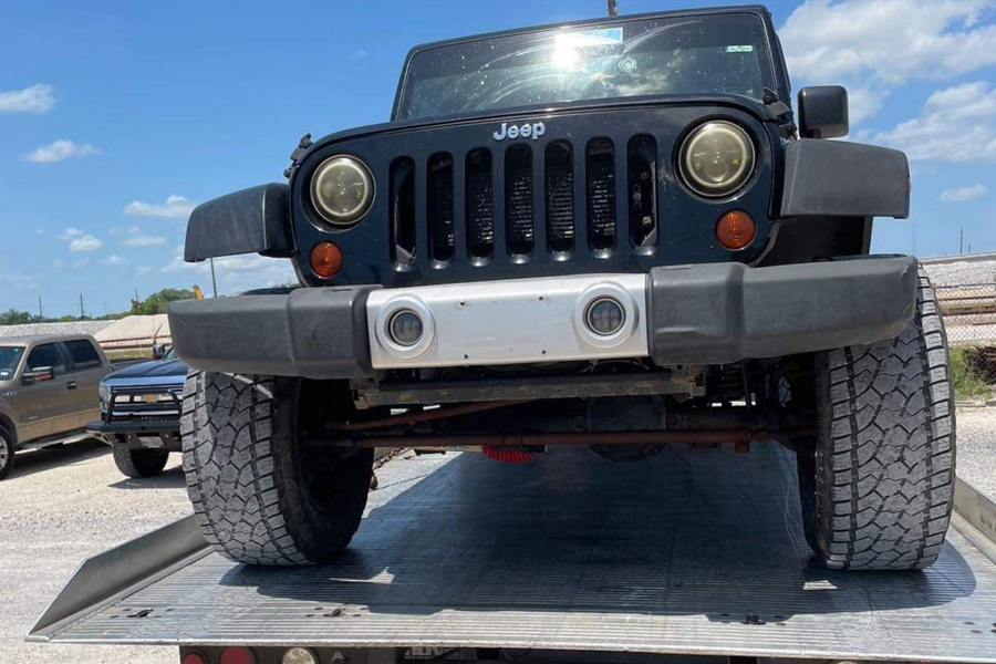 Black Jeep Wrangler on a car trailer, front view in a sunny outdoor lot.