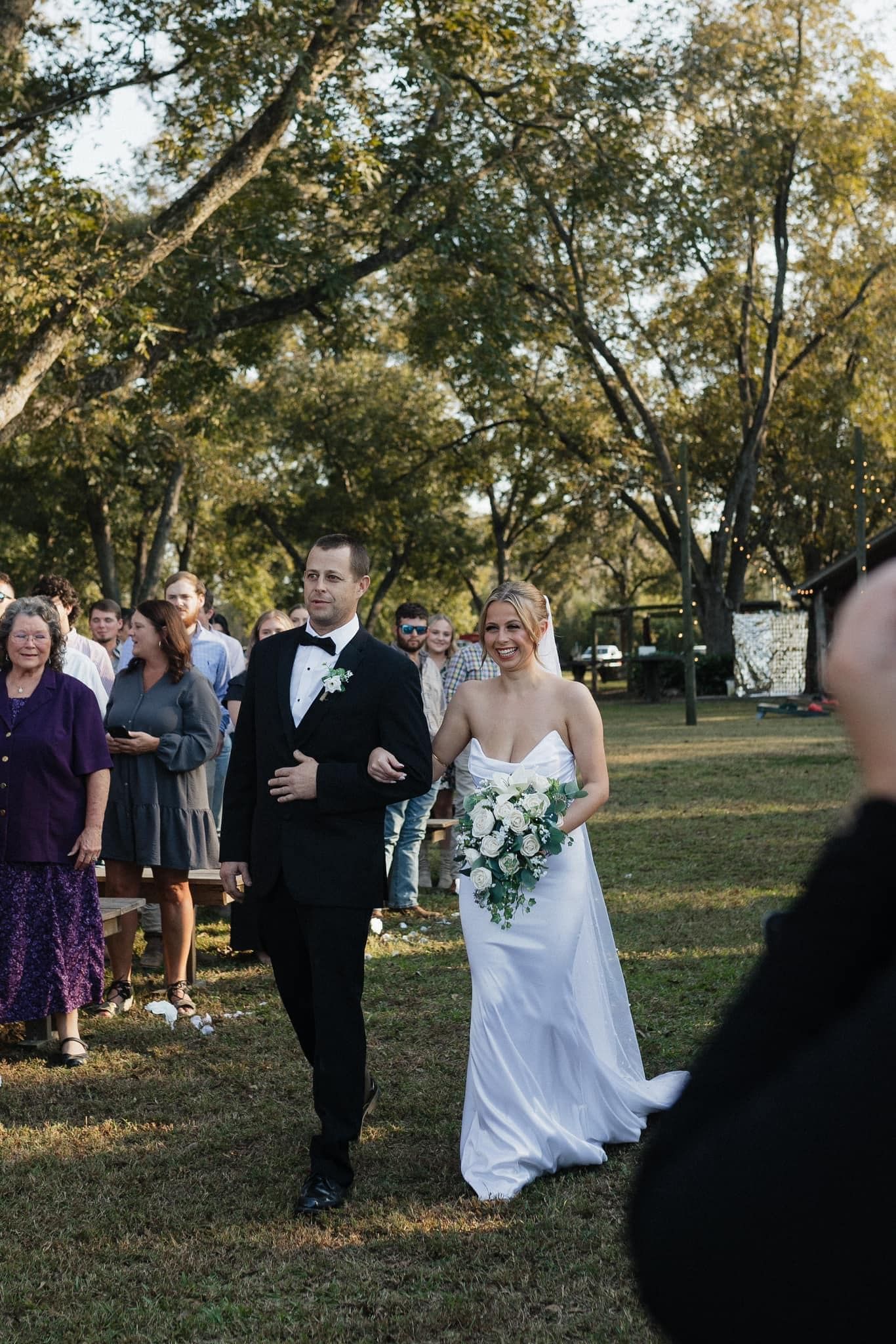 Bride and groom walking outdoors at a wedding, with guests nearby and trees in the background