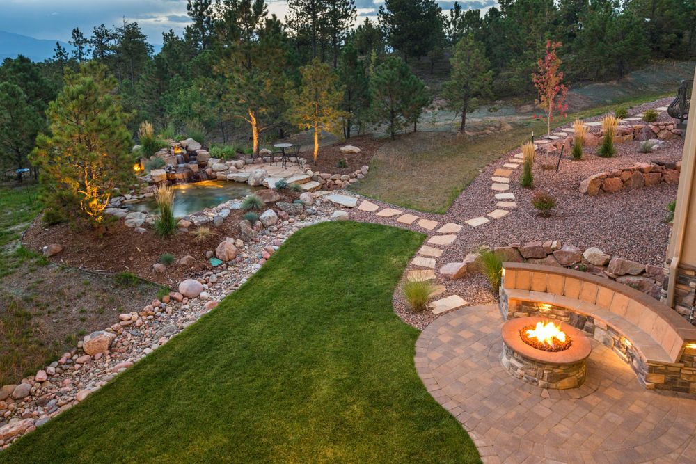 A backyard featuring a stone fire pit with a curved bench, a green lawn, and a small pond, illuminated at dusk.