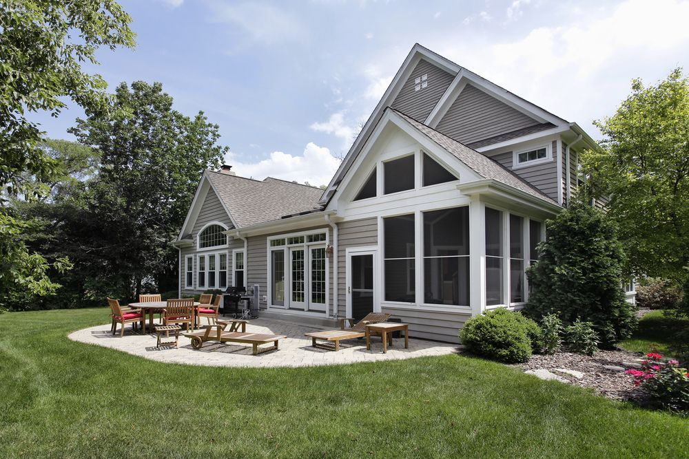 A light gray house with a stone patio, outdoor furniture, and a screened-in porch, surrounded by lush green lawn and trees.