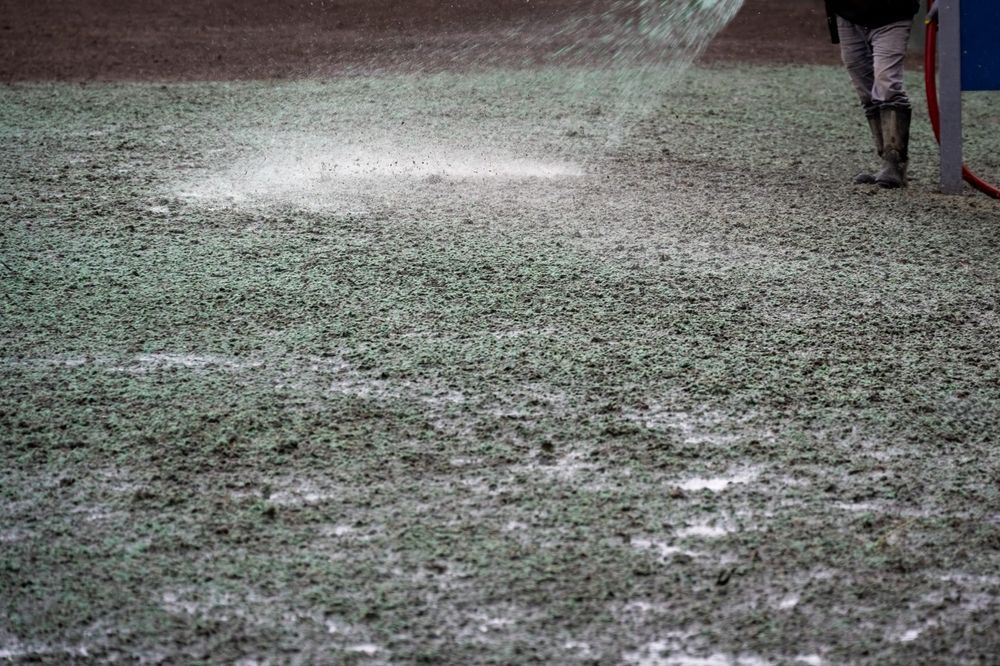 A person wearing boots uses a hose to spray a fine white mist over a patch of grass on a sports field.