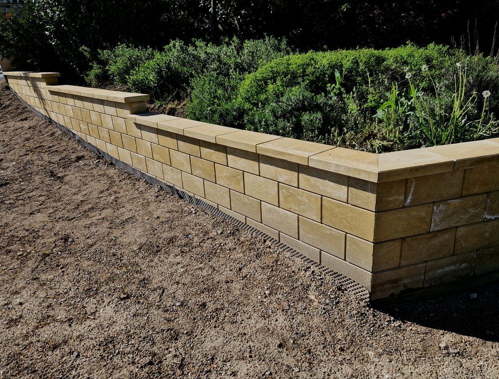 A low, tan stone retaining wall borders a garden bed filled with green plants next to an area of brown dirt.