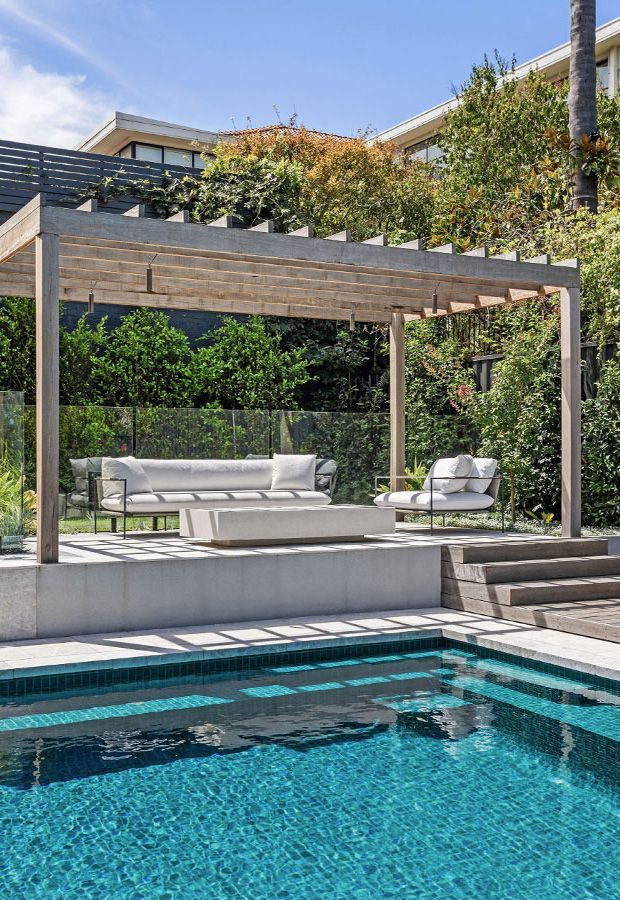 Poolside lounge area with white furniture under a wooden pergola, surrounded by lush greenery.