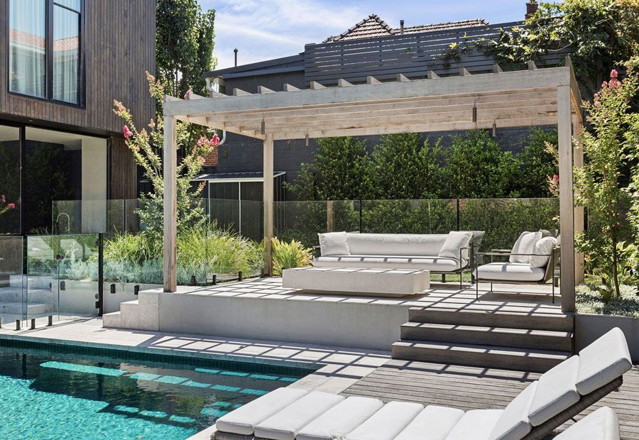 Outdoor lounge area with a pool. Features a white pergola, seating, and lush landscaping.
