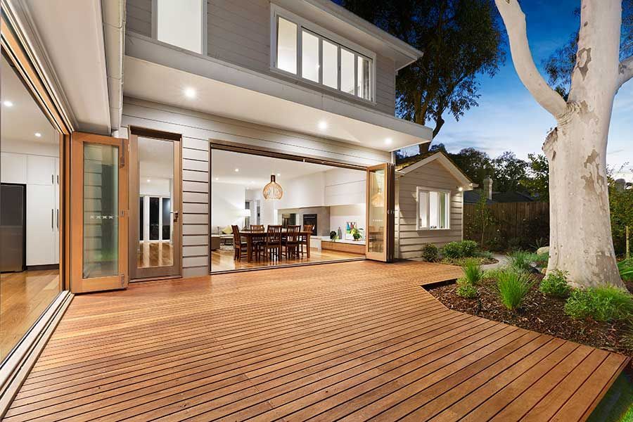 Wooden deck extends from a two-story home with open doors. Backyard with tree and small shed.