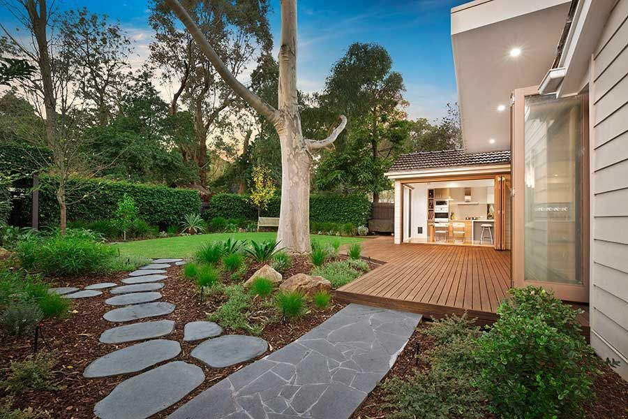 Garden path leads to a wooden deck with a small room open, trees and green grass.
