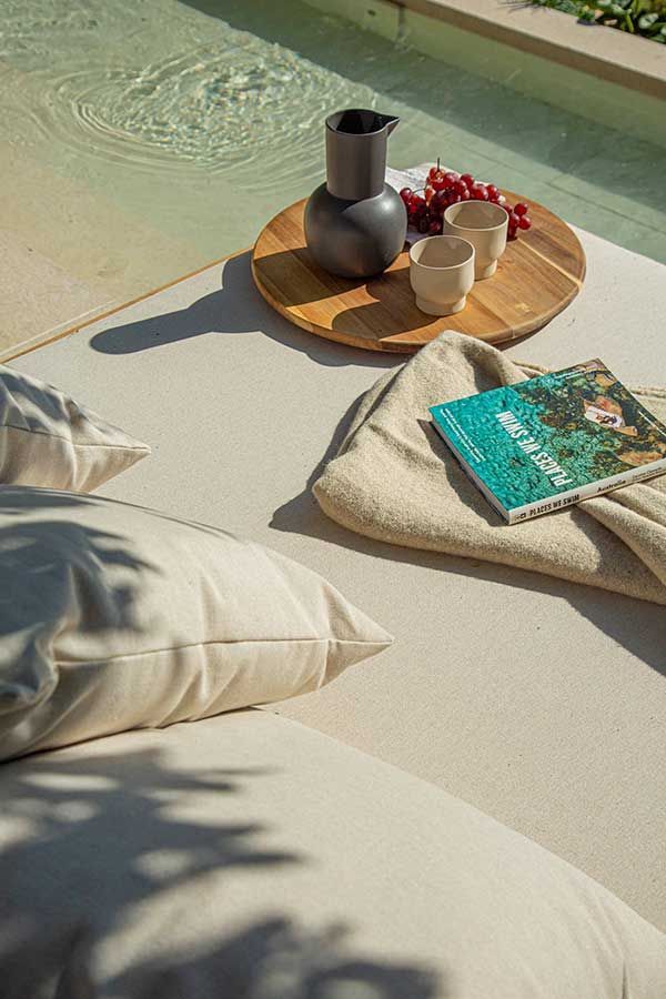 Lounge area by a pool with a tray of drinks and fruit, a book, and pillows.