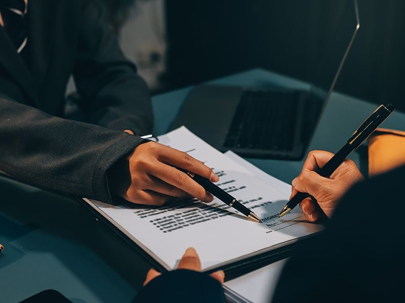 Hands reviewing a document with a pen beside a laptop in a dimly lit office