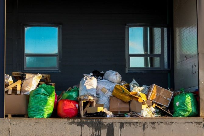 Garbage bags and cardboard boxes piled on a loading dock outside a dark building with two windows