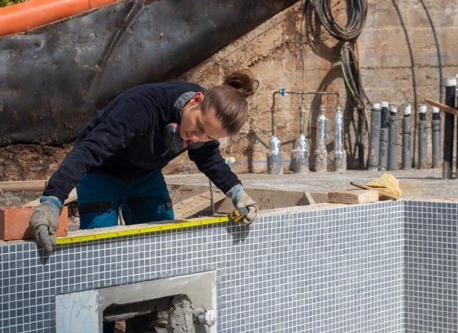 Person measuring the edge of a tiled pool under construction; wearing gloves and a respirator.