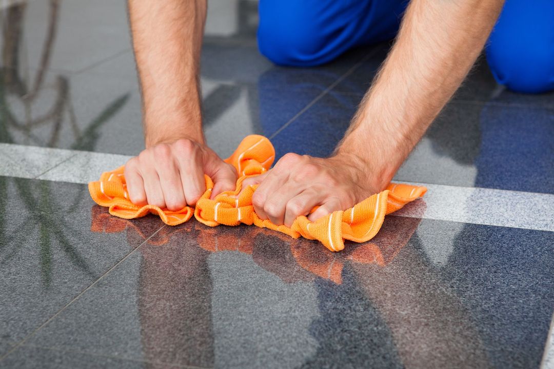 Hands wiping a wet floor with an orange cloth on a gray tiled surface