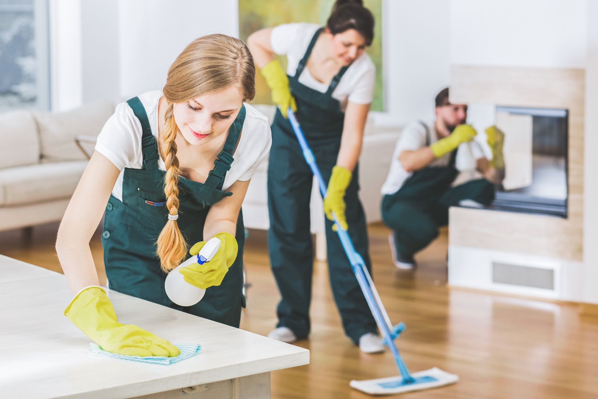 Three people cleaning a bright living room with mops, gloves, and spray bottles.