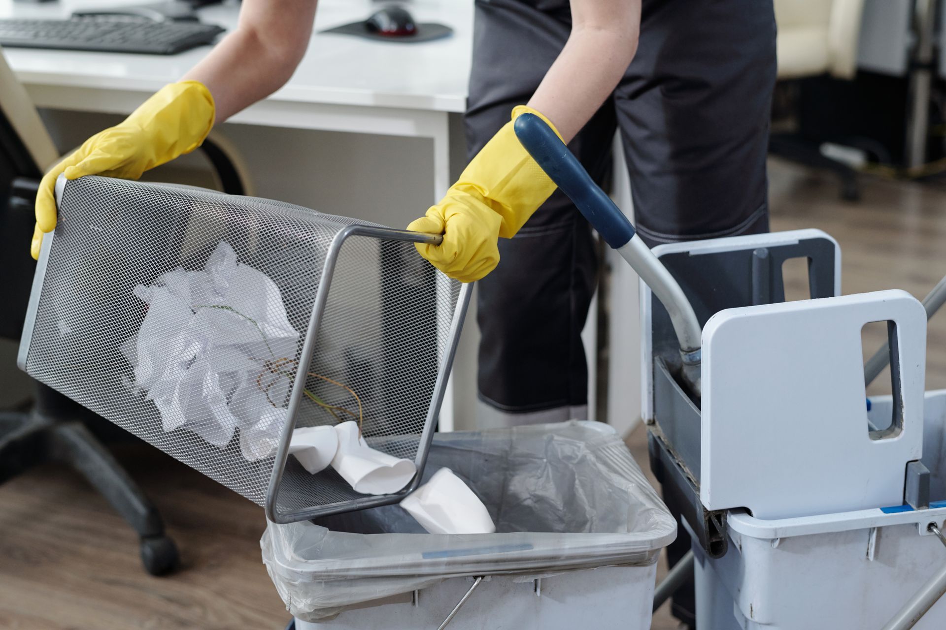 Person in yellow gloves emptying trash into a bin beside a cart in an office or industrial setting
