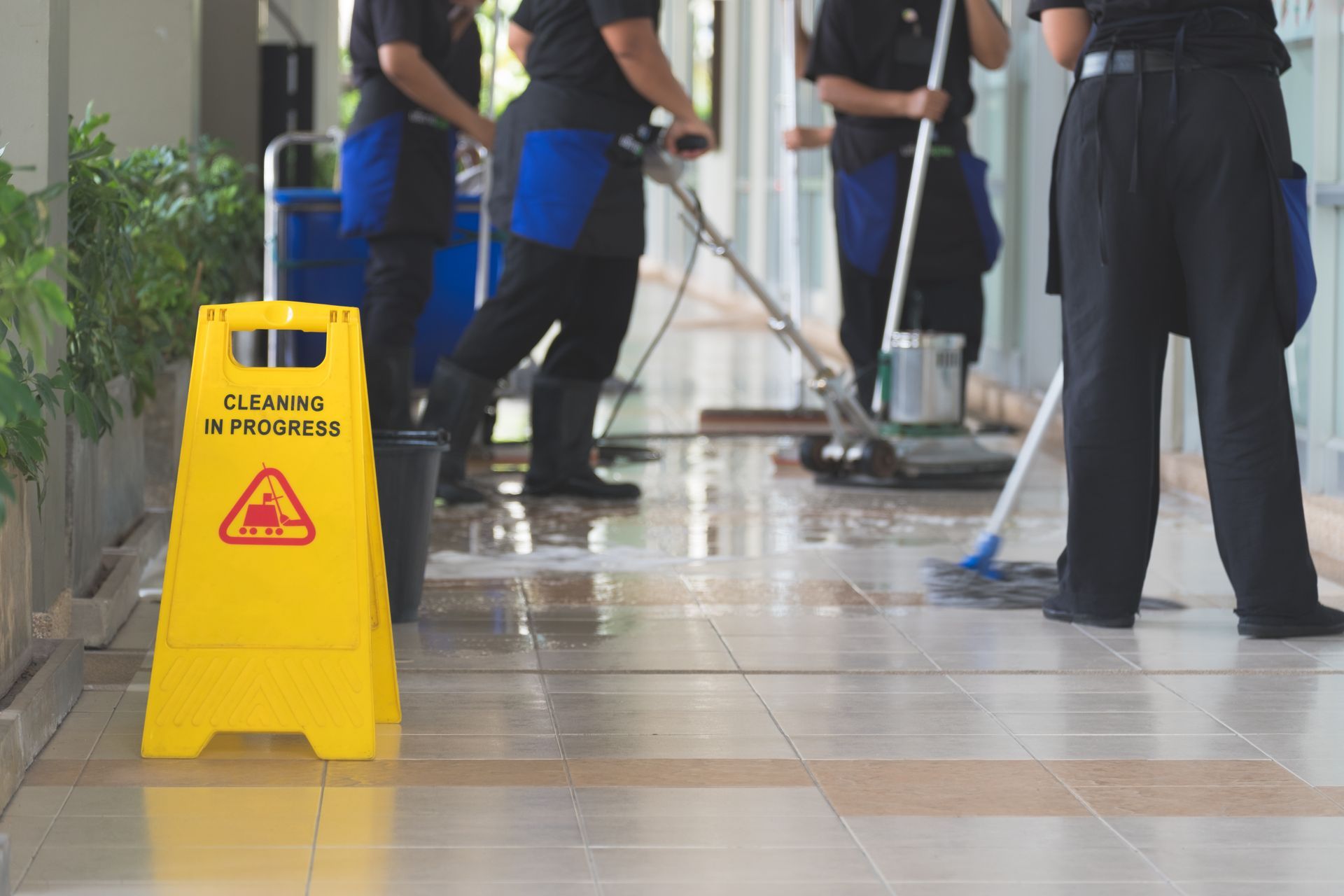Yellow wet-floor sign in a hallway while cleaners mop the floor in the background