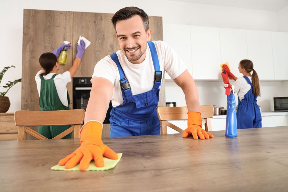 Man in blue overalls cleaning a kitchen countertop with orange gloves, while others clean in the background
