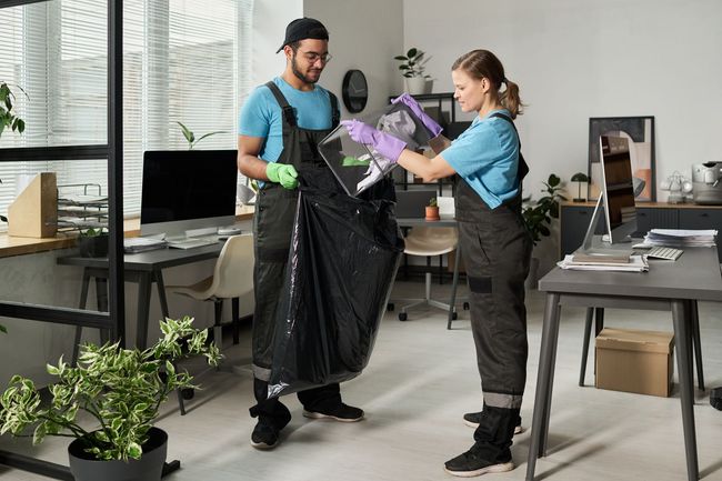 Two workers in blue shirts bag a trash bin in a modern office space.