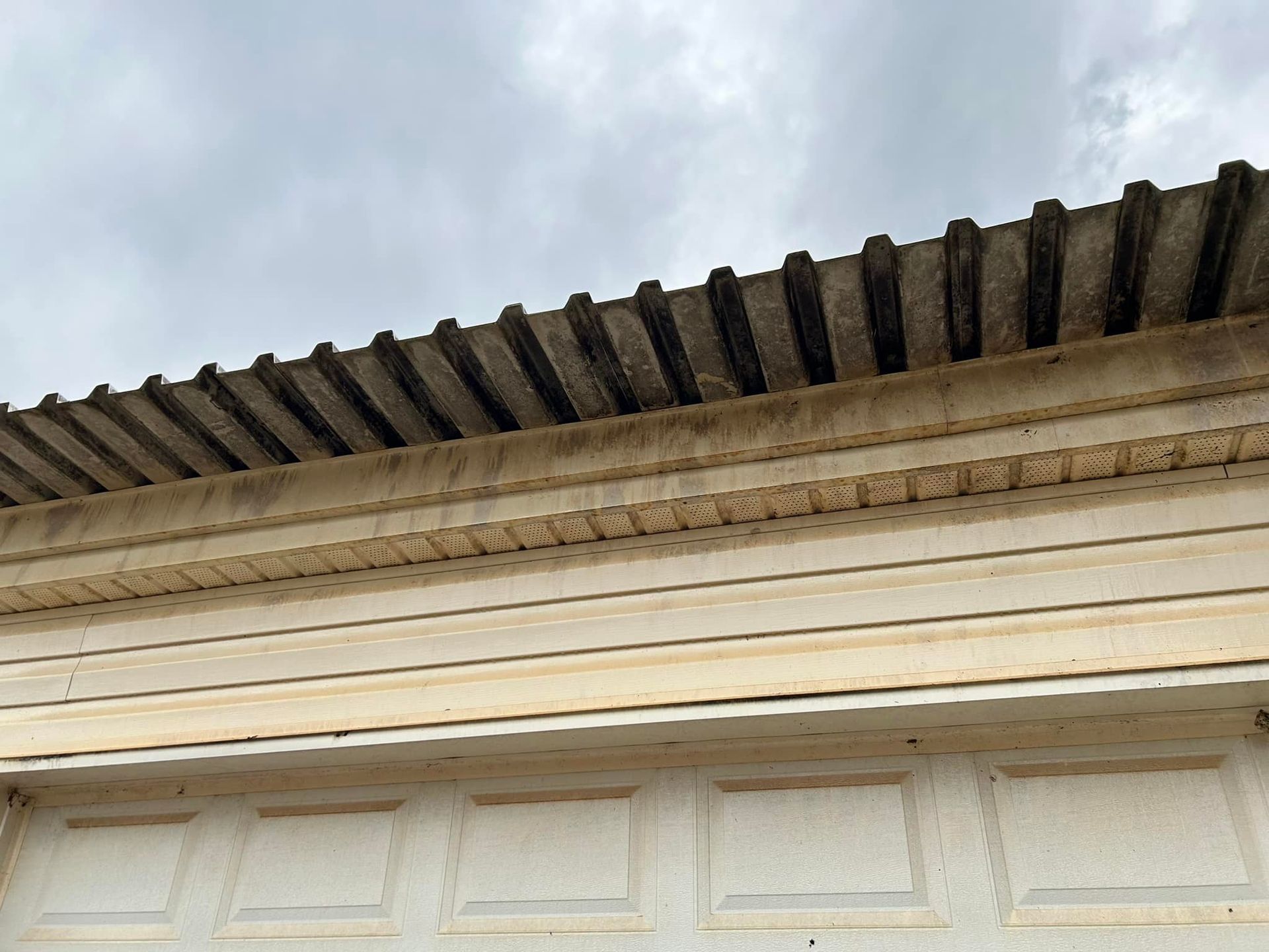 Upward view of a cream-colored building facade with ornate trim and a tiled roof edge against a cloudy sky