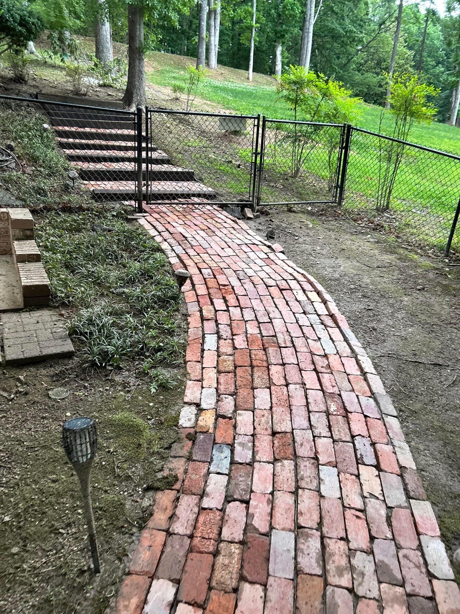 Curved red brick walkway up a hillside, bordered by black railings and greenery in a wooded yard