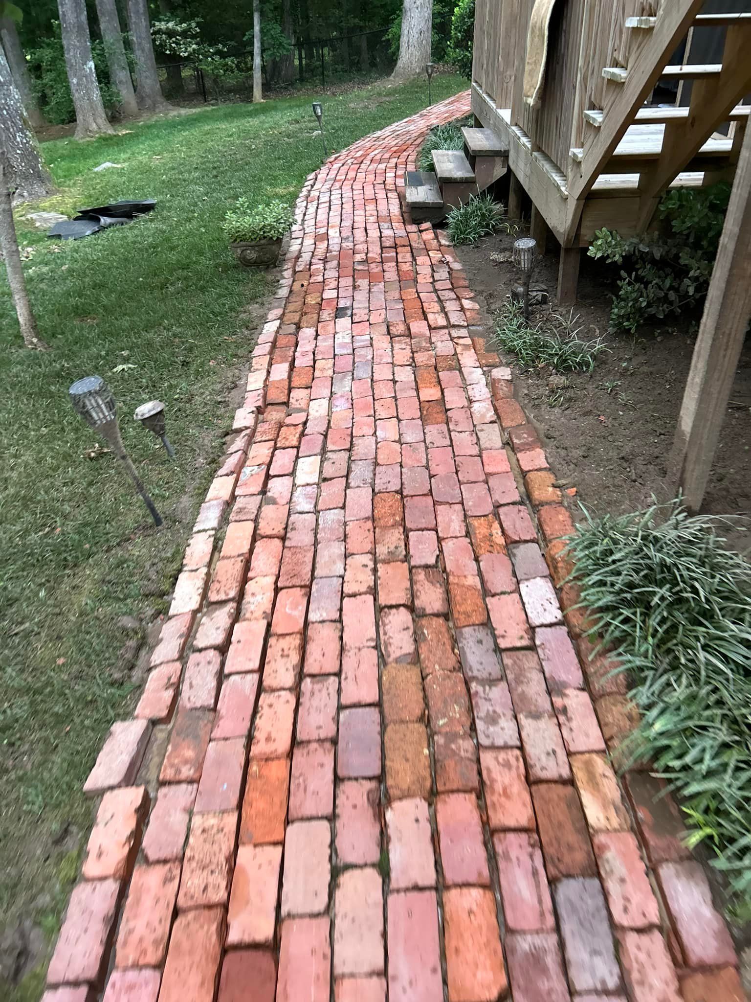Brick pathway beside a wooden deck, bordered by grass and trees in a yard.