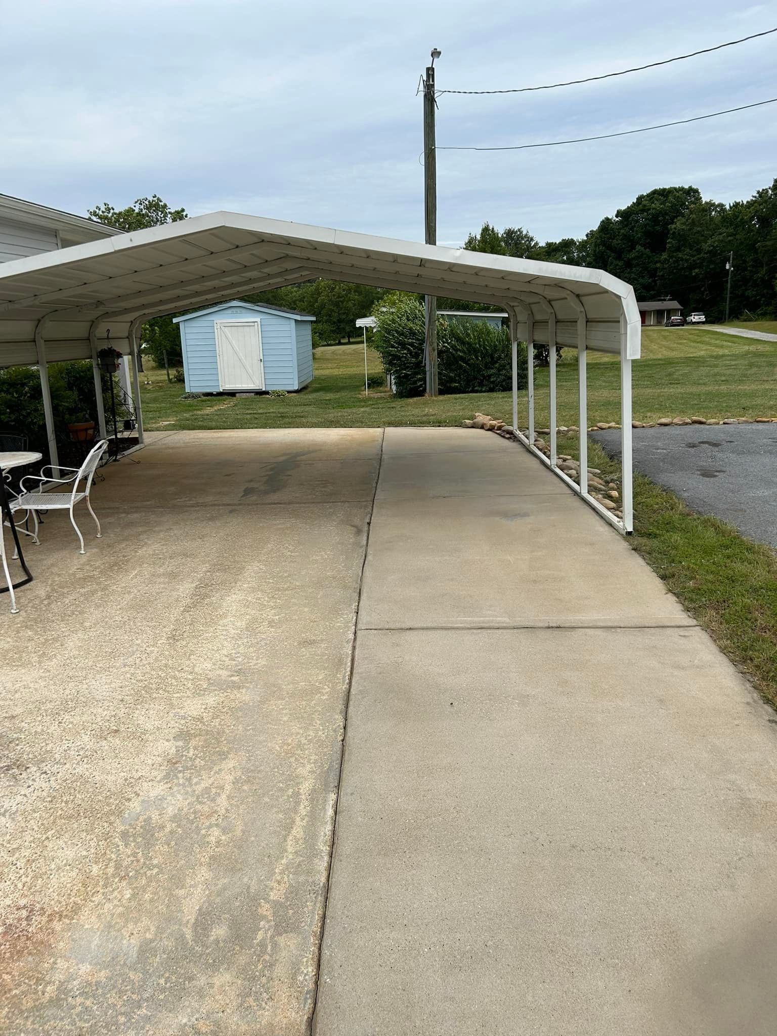 Covered driveway/carport beside a concrete path, with a road and trees in the background.