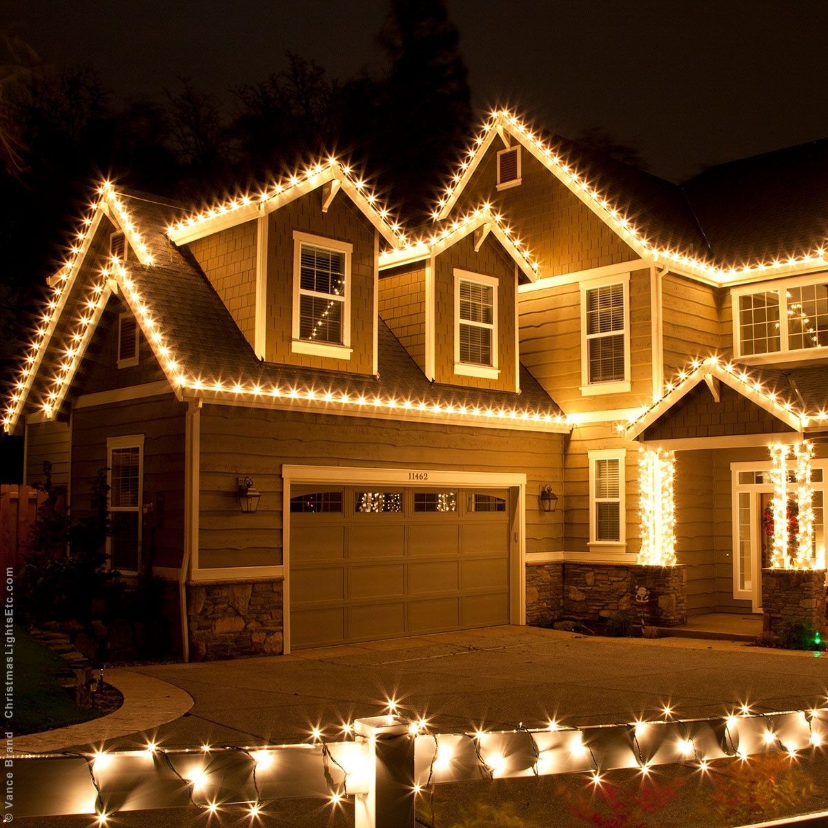 Two-story house outlined with warm white Christmas lights at night, with lit garland along the front yard