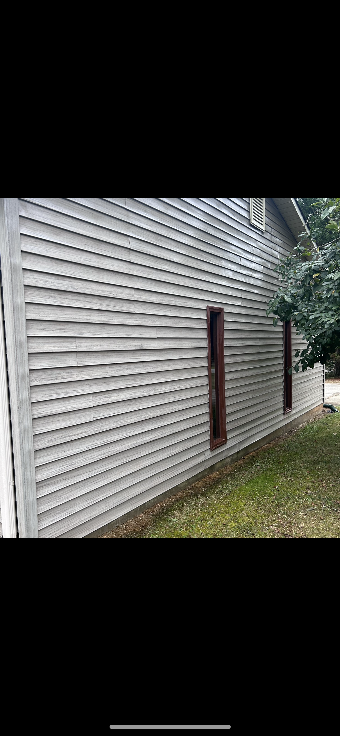 A weathered, gray-sided building with dark wooden posts; grass and trees in the background.